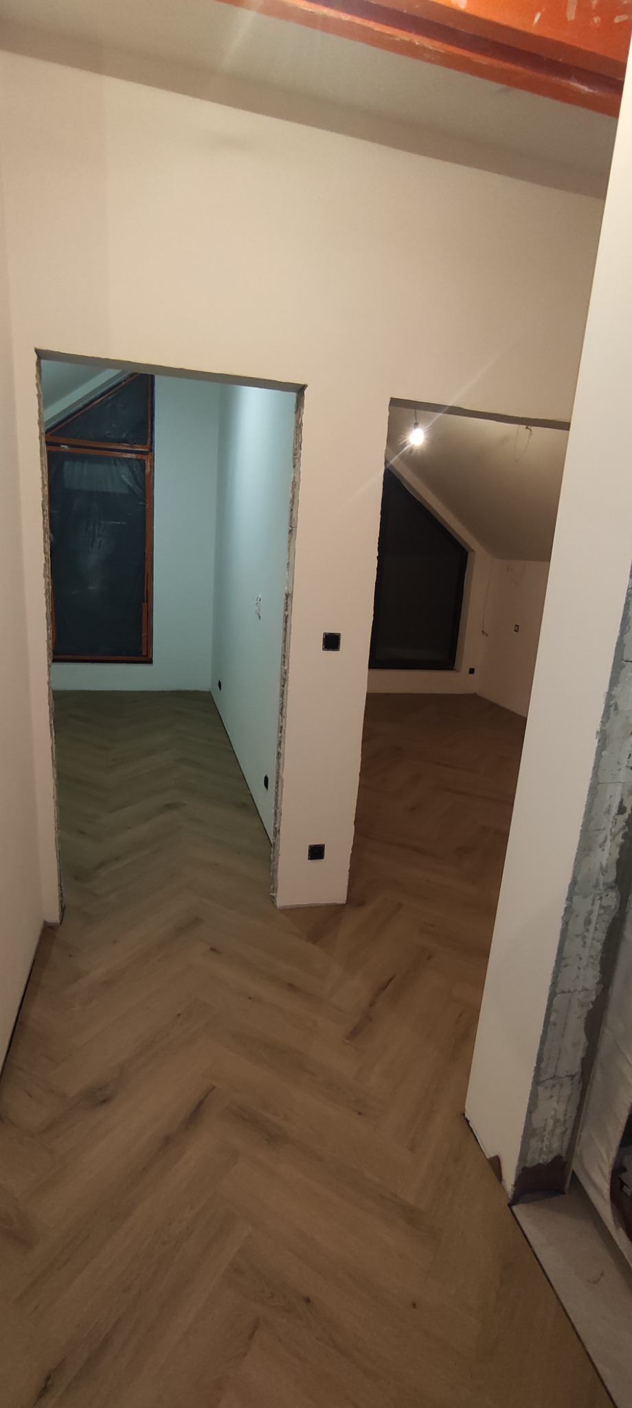 Ukończone poddasze z ułożoną podłogą w jodełkę, widoczne dwa otwory drzwiowe i okno dachowe z czarną folią ochronną.