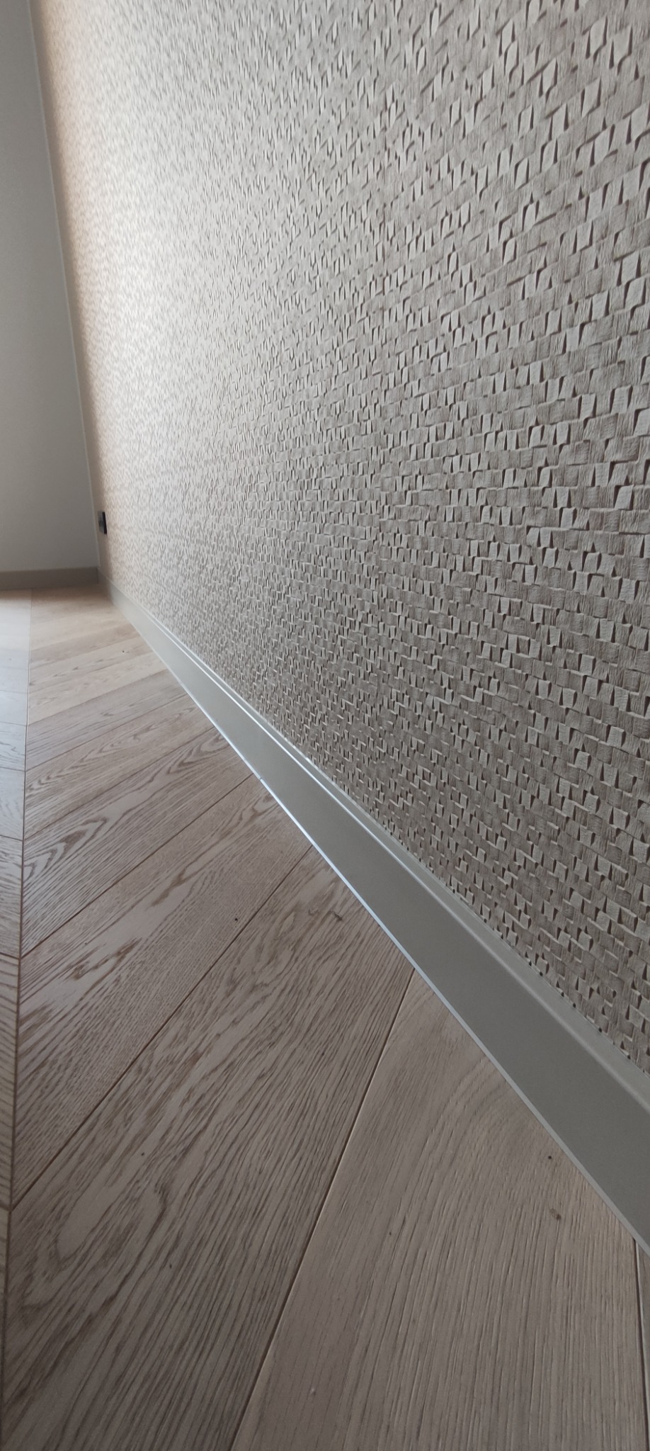 Jasne panele podłogowe ułożone w jodełkę, zakończone białą listwą przypodłogową, na ścianie dekoracyjna tapeta z geometrycznym wzorem.