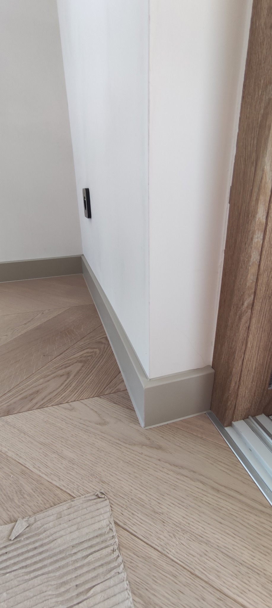 Szczegółowe ujęcie nowo położonej podłogi z paneli w jodełkę, z widocznymi szarymi listwami przypodłogowymi i framugą drzwi po prawej stronie.