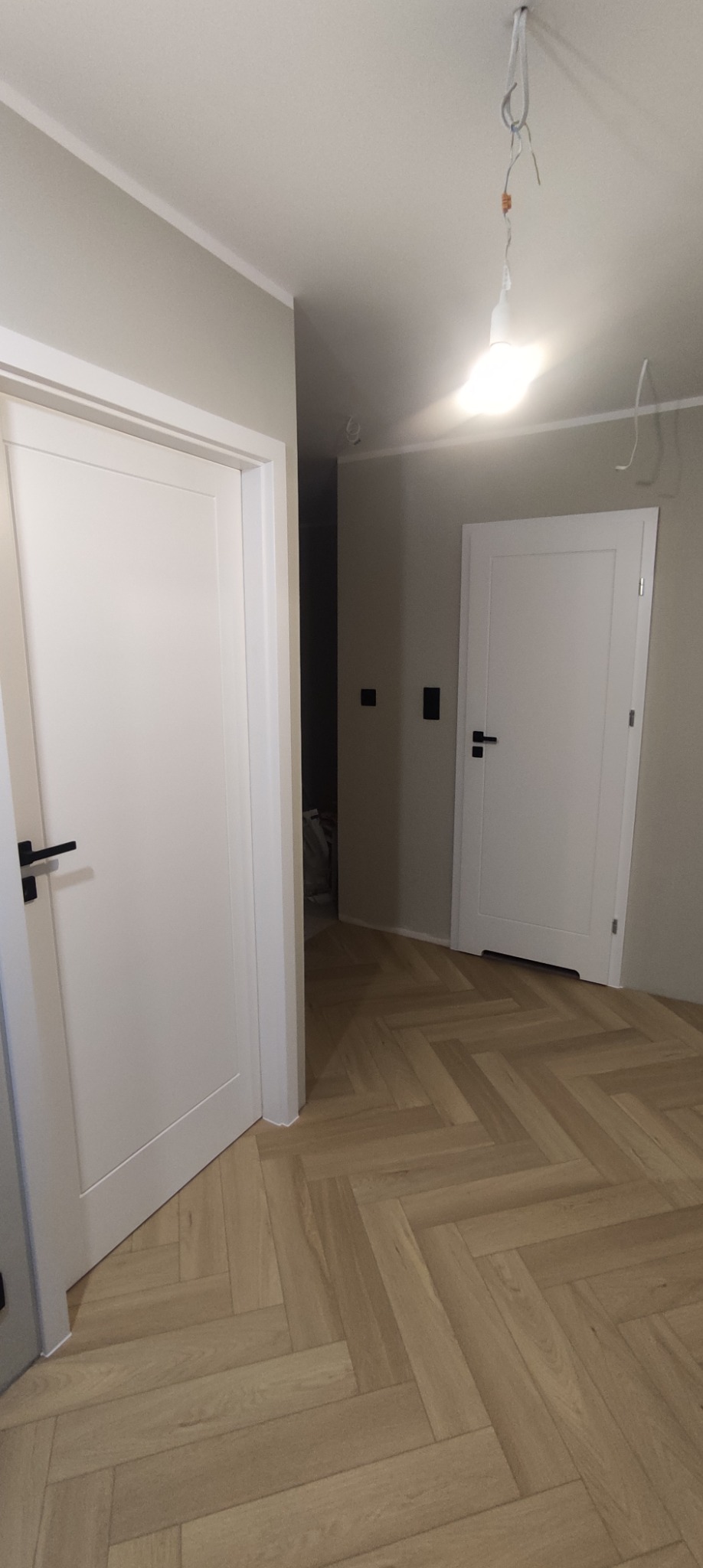 Wnętrze z jasną podłogą ułożoną w jodełkę i dwoma białymi drzwiami z czarnymi klamkami, widoczna surowa żarówka na kablu.