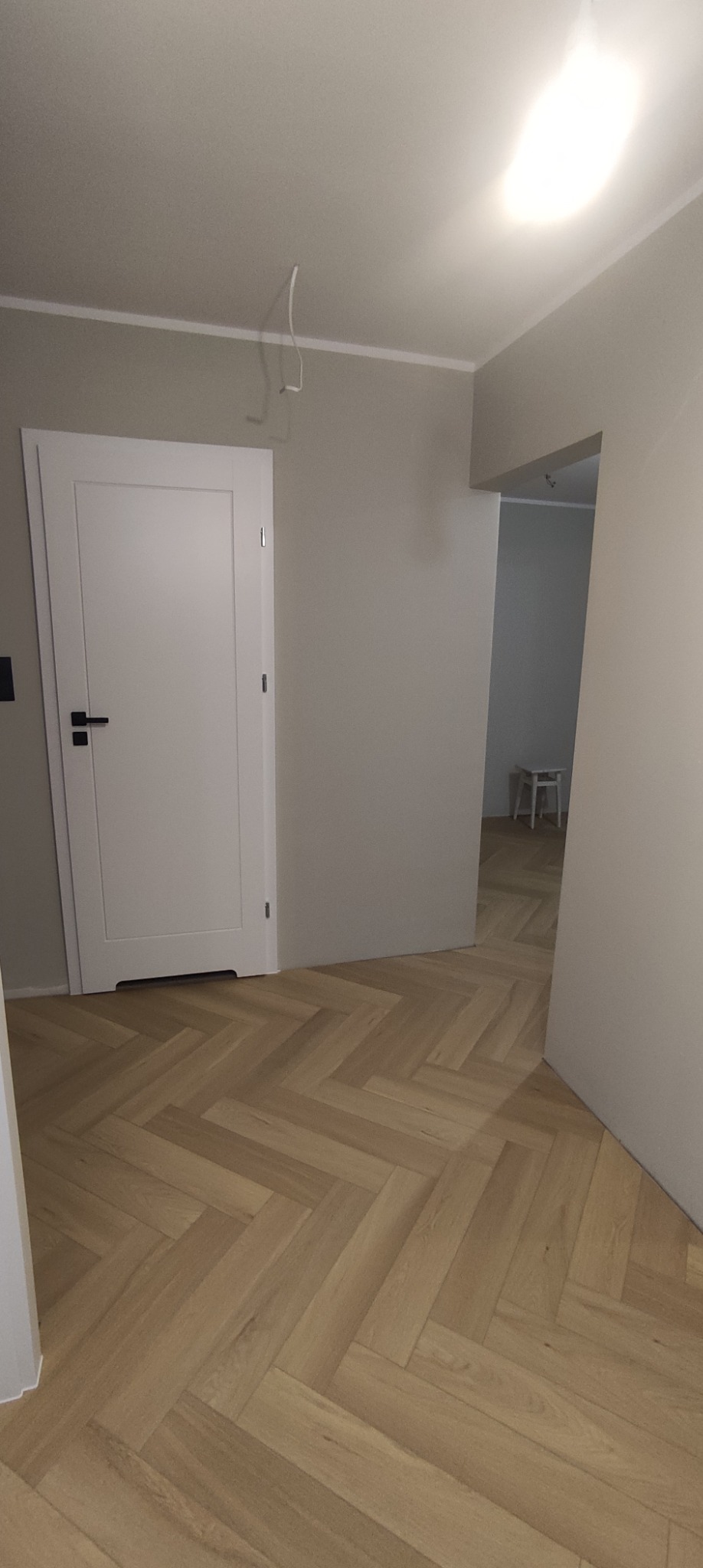 Wnętrze z jasną podłogą ułożoną w jodełkę, białe drzwi z czarną klamką, szare ściany i widok na kolejne pomieszczenie z białym stołkiem.