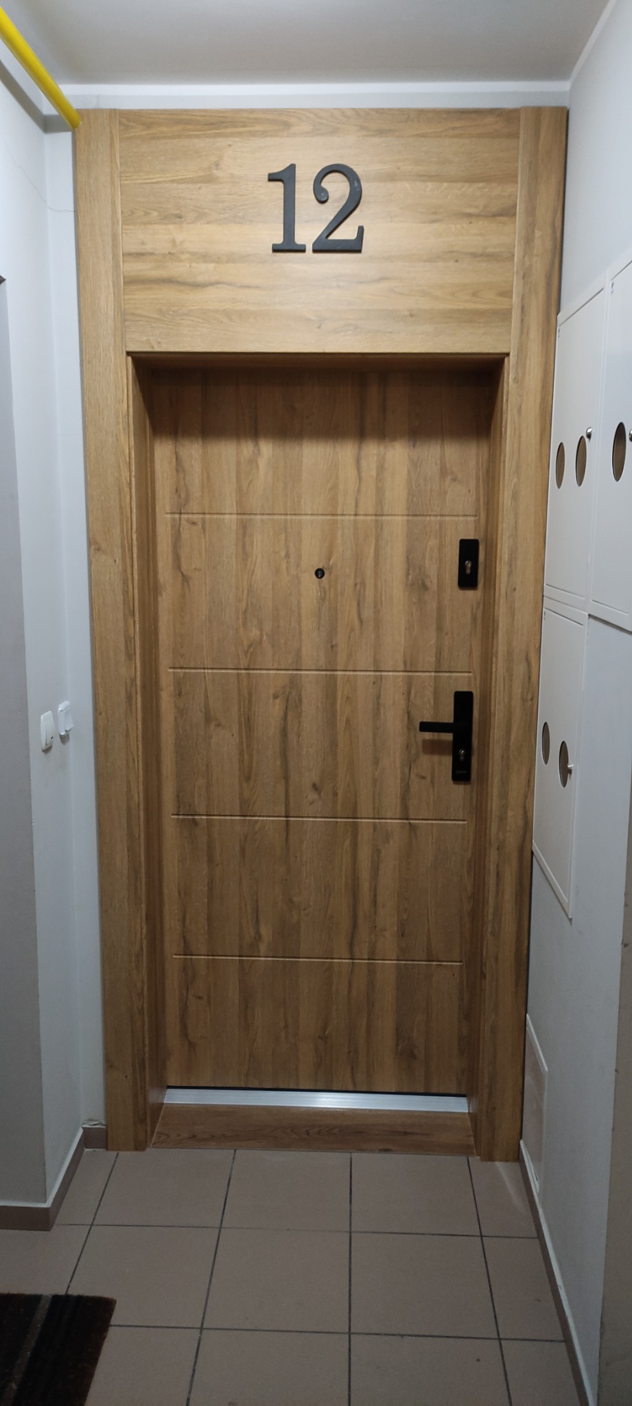 Drewniane drzwi wejściowe do mieszkania z numerem 12, czarną klamką i wizjerem, zamontowane w bloku mieszkalnym.