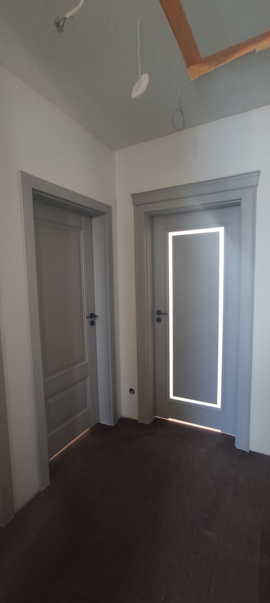 Dwa szare, wewnętrzne drzwi z czarnymi klamkami w białym pomieszczeniu z ciemną podłogą. Jeden z drzwi posiada podłużne przeszklenie.