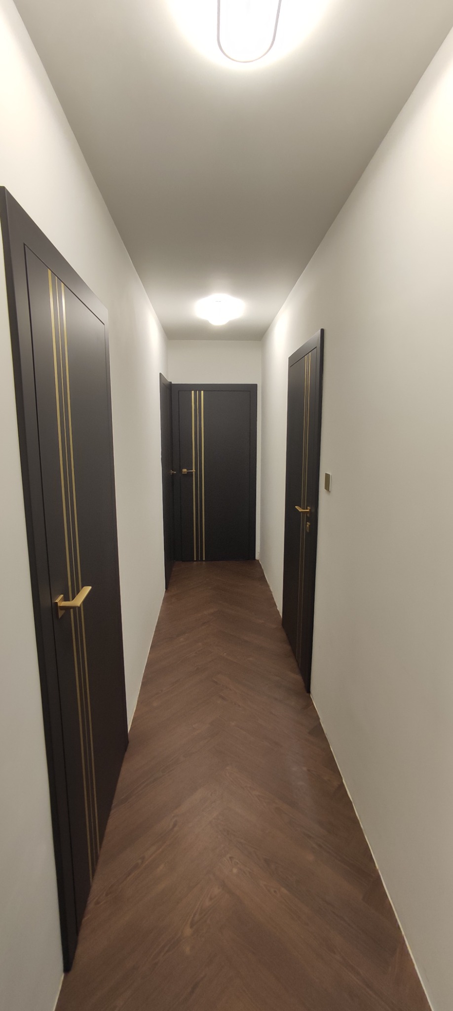 Korytarz z brązową podłogą ułożoną w jodełkę i trzema czarnymi drzwiami z złotymi detalami. Dwa źródła światła sufitowego.