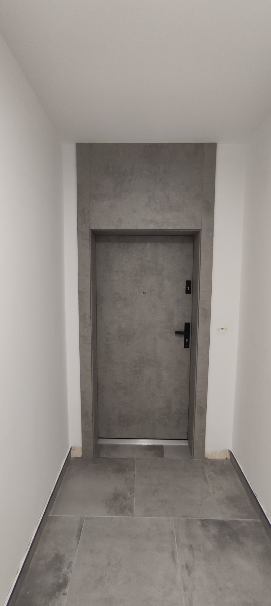 Szare drzwi wejściowe z czarną klamką i wizjerem, otoczone szarą, kamienną ramą w białym korytarzu z szarą podłogą z płytek.