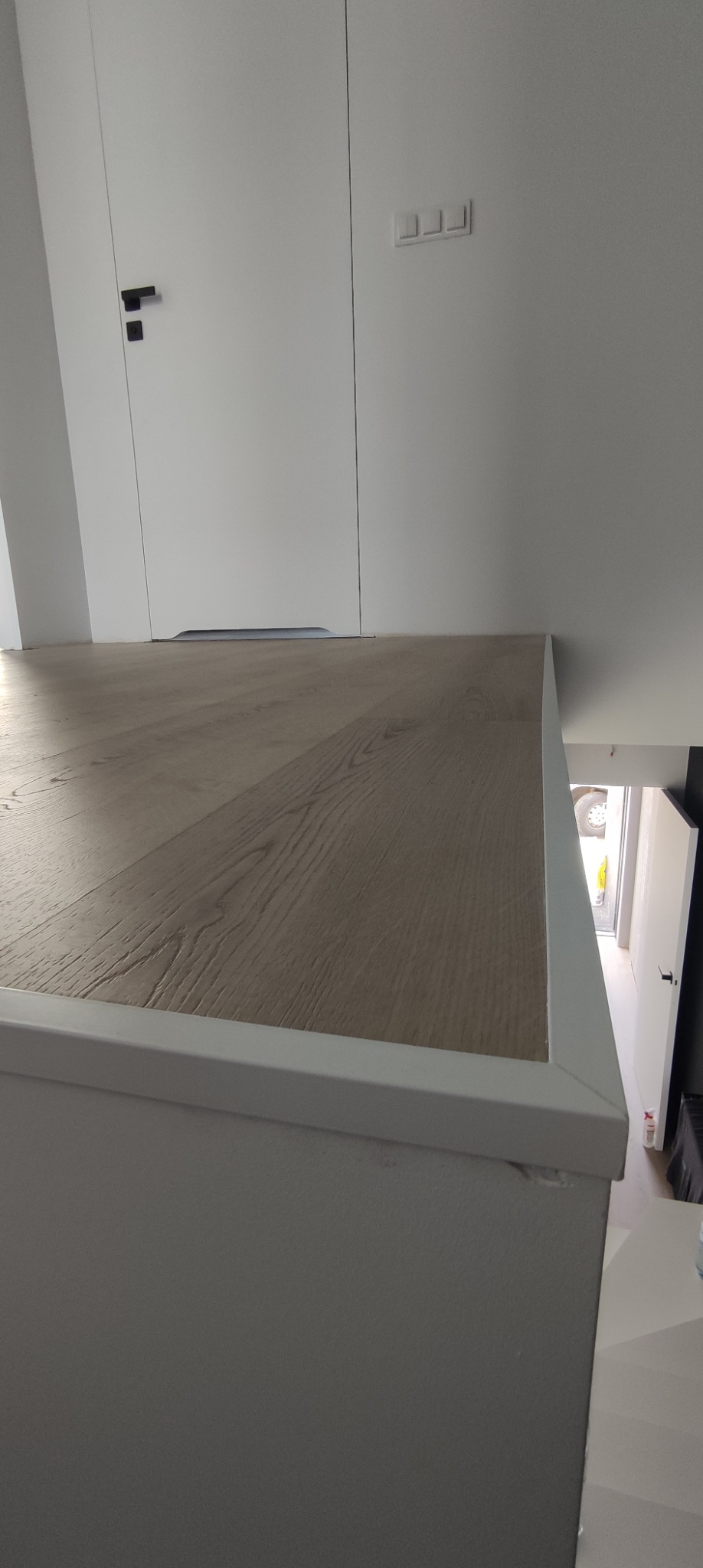 Fragment podłogi z panelami laminowanymi w odcieniach beżu i szarości, zakończonej białą listwą przypodłogową, widoczna z góry, z białymi drzwiami w tle