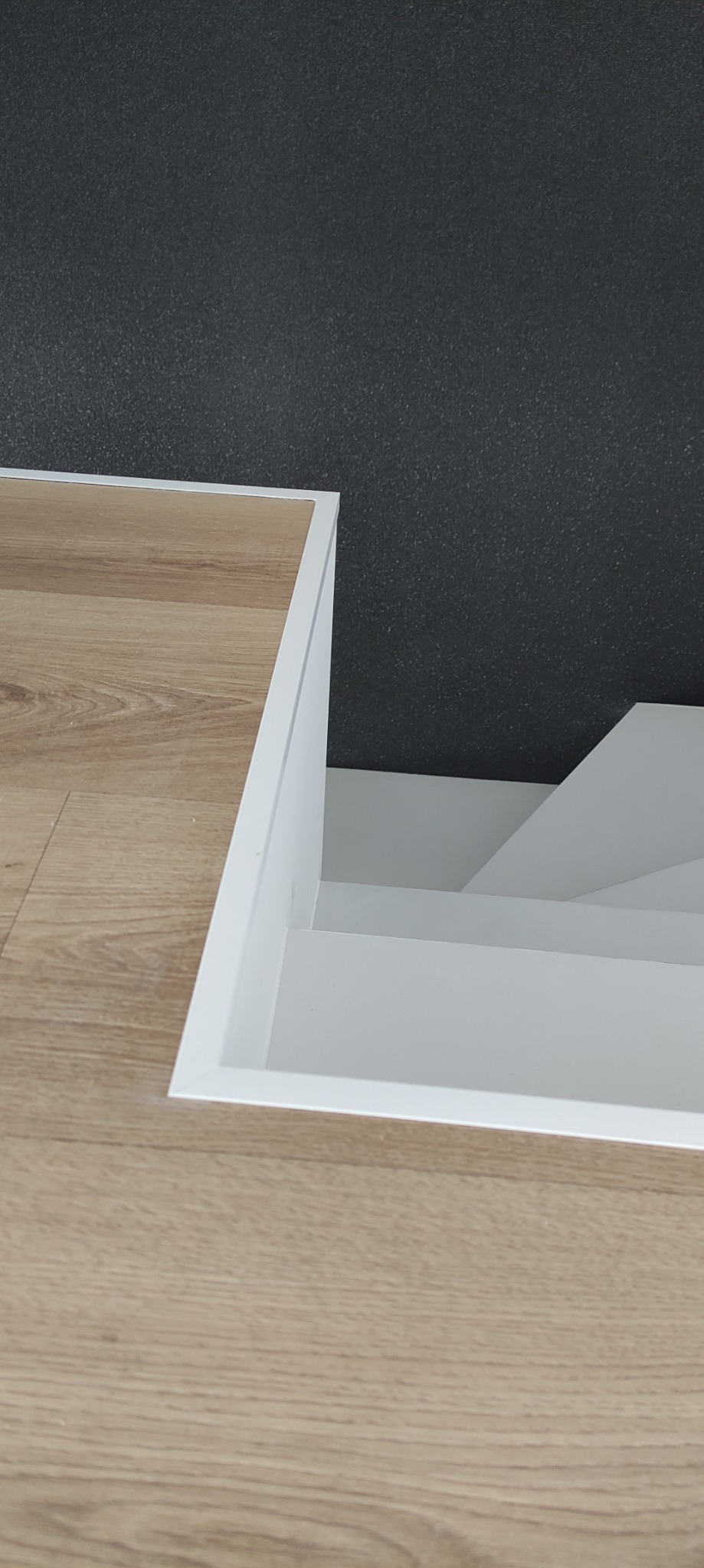 Fragment podłogi z paneli imitujących drewno, z widocznym wejściem do piwnicy lub schowka wykończonym białymi listwami.