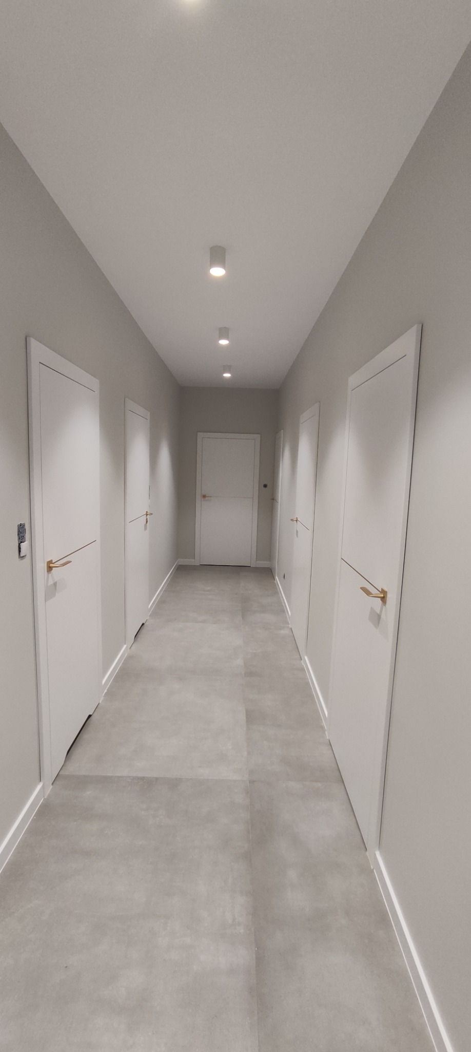Długi korytarz z szarymi płytkami podłogowymi i białymi drzwiami wewnętrznymi z poziomymi, złotymi klamkami. Sufit z wbudowanymi, punktowymi lampami.