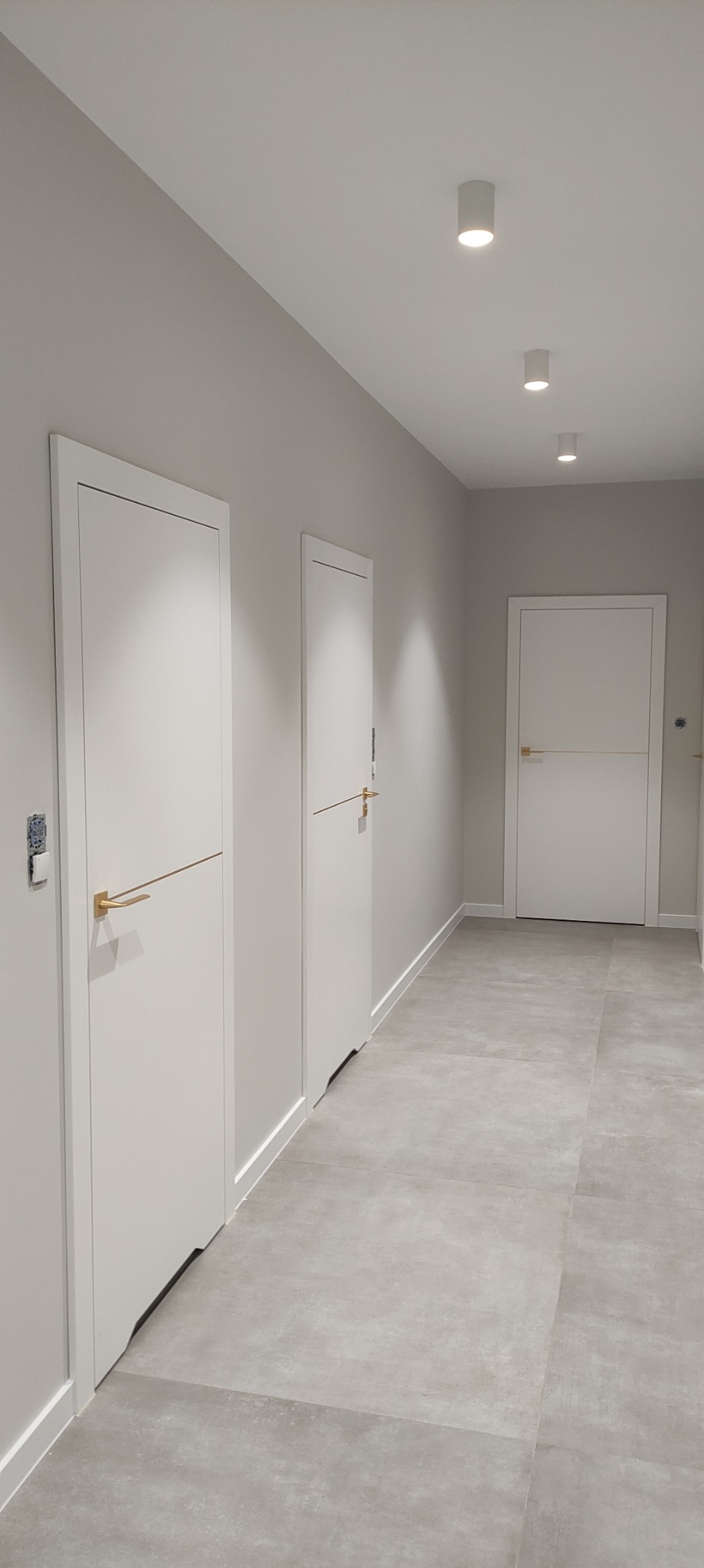 Korytarz z trzema białymi, minimalistycznymi drzwiami z poziomymi, złotymi klamkami, szara podłoga z płytek, oświetlenie sufitowe punktowe.