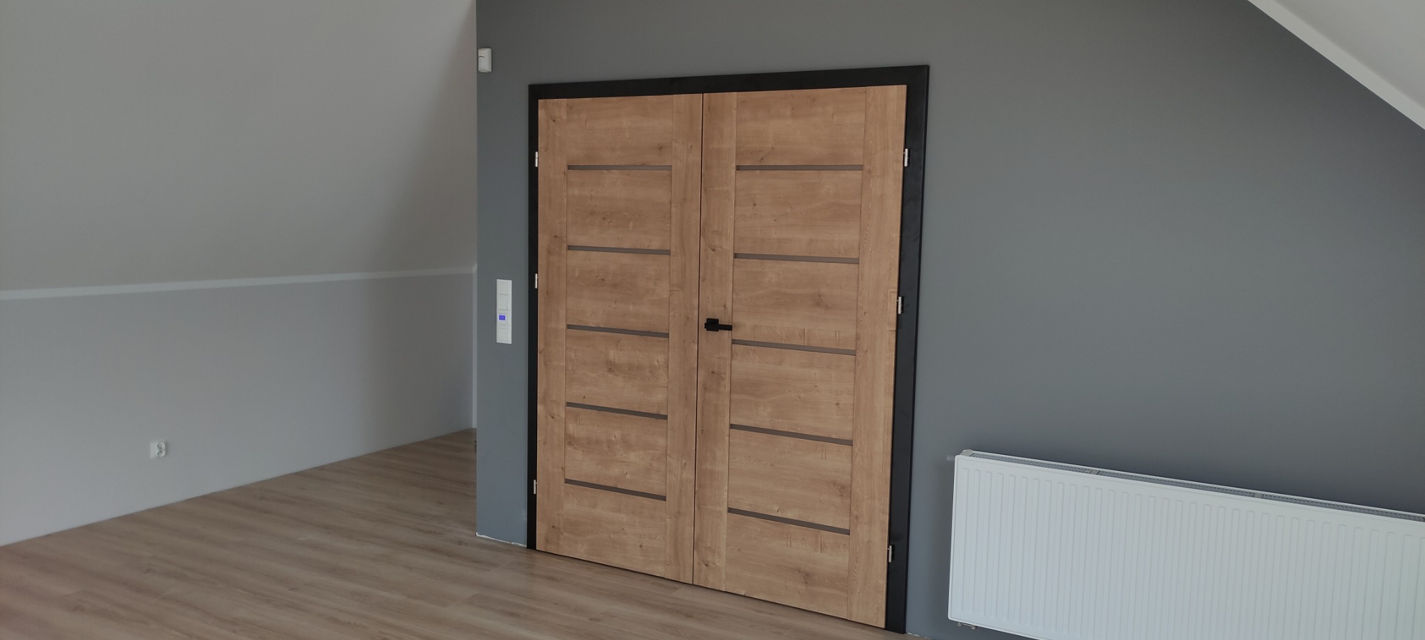 Dwuskrzydłowe, drewniane drzwi wewnętrzne z czarną ościeżnicą, zamontowane w pokoju na poddaszu, widoczne z perspektywy podłogi wyłożonej panelami.