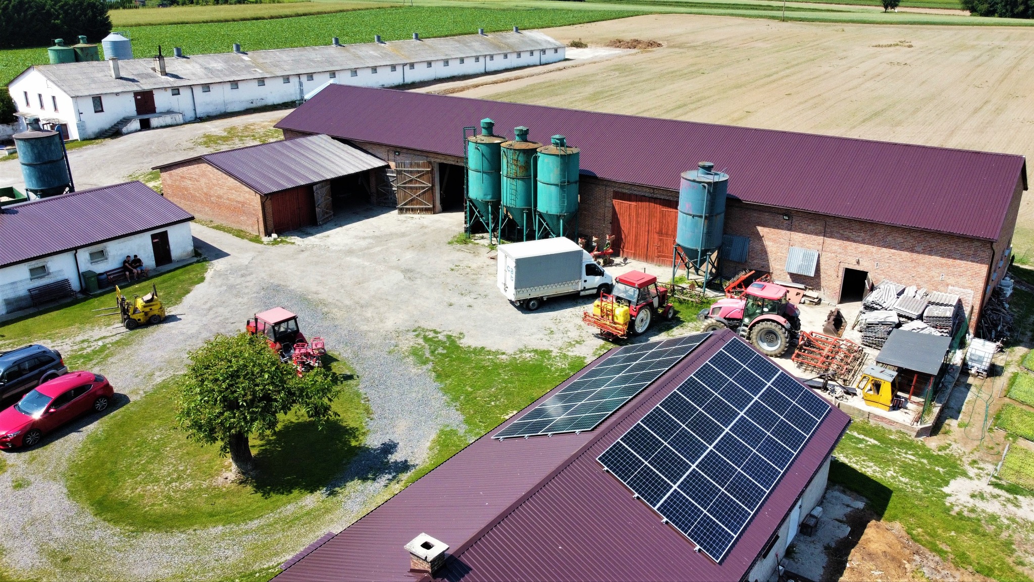 Widok z góry na farmę z zainstalowanymi panelami słonecznymi na dachu jednego z budynków, obok stoją traktory i maszyny rolnicze, w tle budynki gospodarcze i pola uprawne.