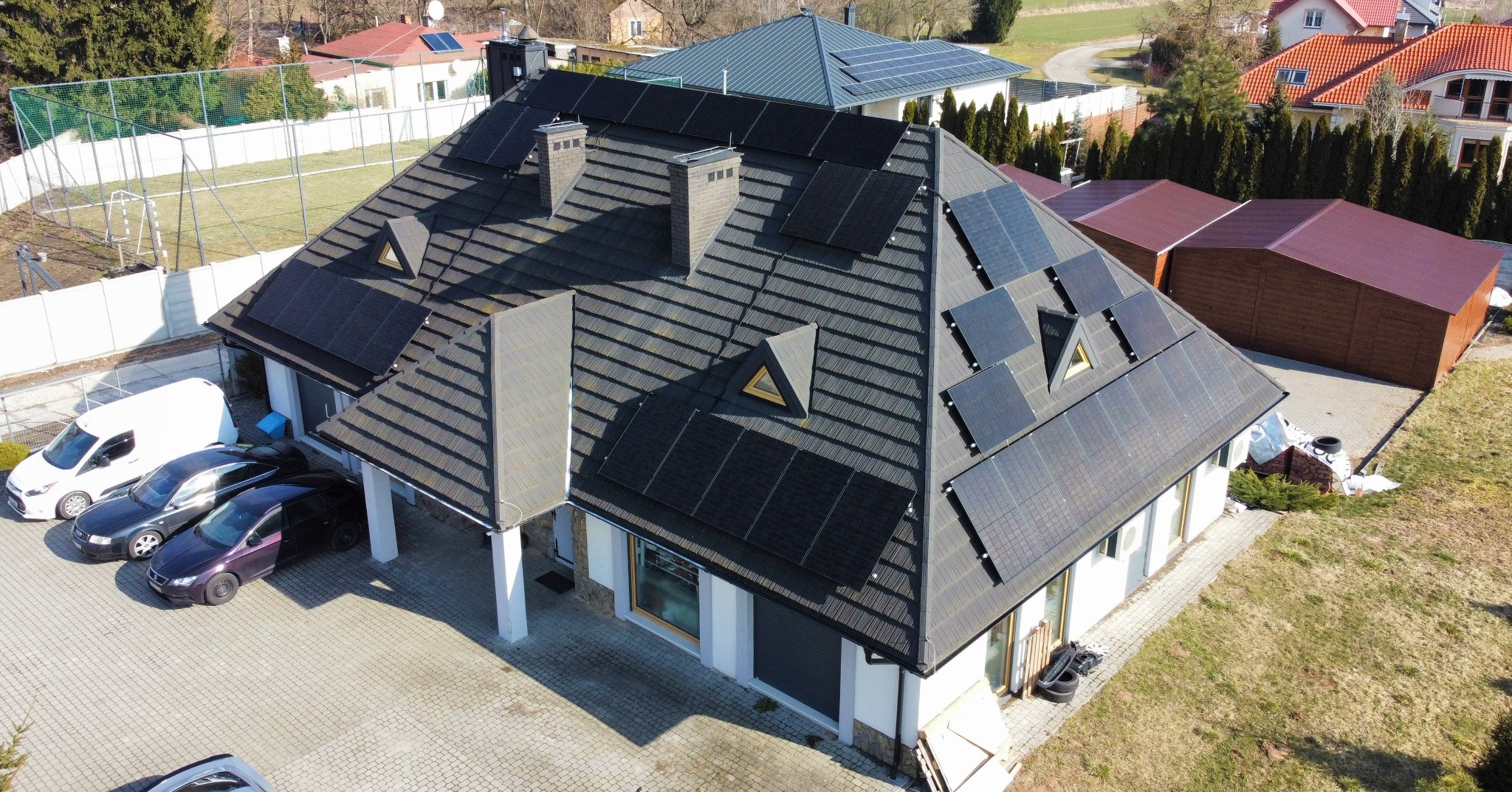 Dom jednorodzinny z panelami słonecznymi na dachu z dachówki ceramicznej, widok z lotu ptaka na podjazd z zaparkowanymi samochodami i otoczenie budynku.