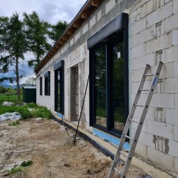 Perfect Windows Glazing Excellence - Surowy stan budynku z białego bloczka z zamontowanymi czarnymi oknami z roletami; widoczna drabina oparta o ścianę, teren wokół budynku jeszcze niezagospodarowany.