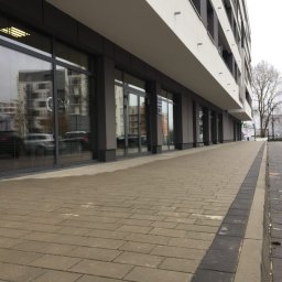 Perfect Windows Glazing Excellence - Fasada nowoczesnego budynku biurowego z dużymi, przeszklonymi witrynami i szarymi ramami okiennymi, odbijającymi otoczenie, widok z poziomu chodnika wyłożonego kostką.