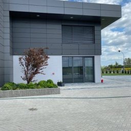 Perfect Windows Glazing Excellence - Nowoczesny budynek biurowy z szarą elewacją i aluminiowymi oknami żaluzjowymi, przeszklone wejście, bruk na pierwszym planie, rabata z zielenią i drzewkiem.