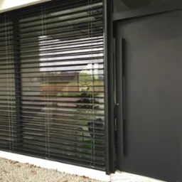 Nowoczesne okno z czarnymi żaluzjami poziomymi, widok z zewnątrz, obok drzwi wejściowe w kolorze antracytowym, jasne otoczenie budynku.