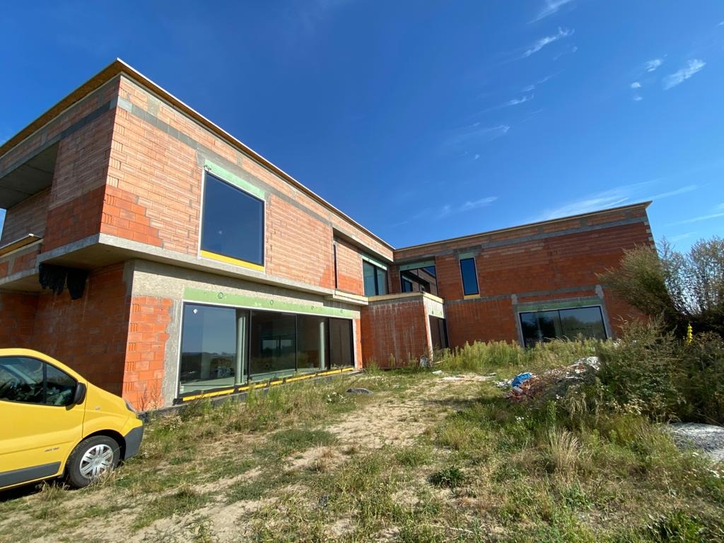 Dwukondygnacyjny budynek w trakcie budowy z wstawionymi oknami z czarnymi ramami, widoczny żółty samochód dostawczy i zarośnięty teren wokół.