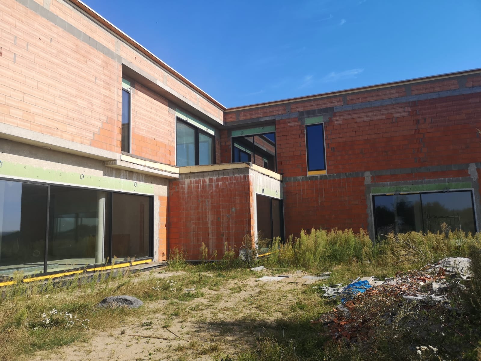 Surowy stan budynku w trakcie montażu okien, widoczne cegły, wstawione ramy okienne z szybami i materiały izolacyjne na zewnątrz, trawa i gruz na pierwszym planie.