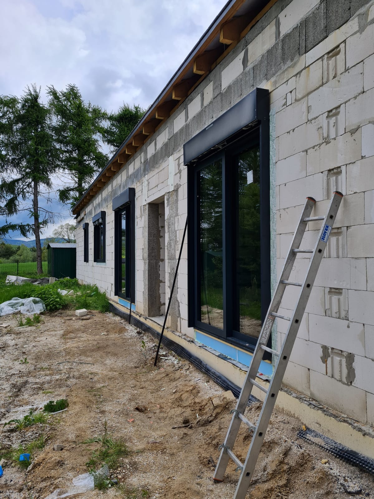 Surowy stan budynku z białego bloczka z zamontowanymi czarnymi oknami z roletami; widoczna drabina oparta o ścianę, teren wokół budynku jeszcze niezagospodarowany.
