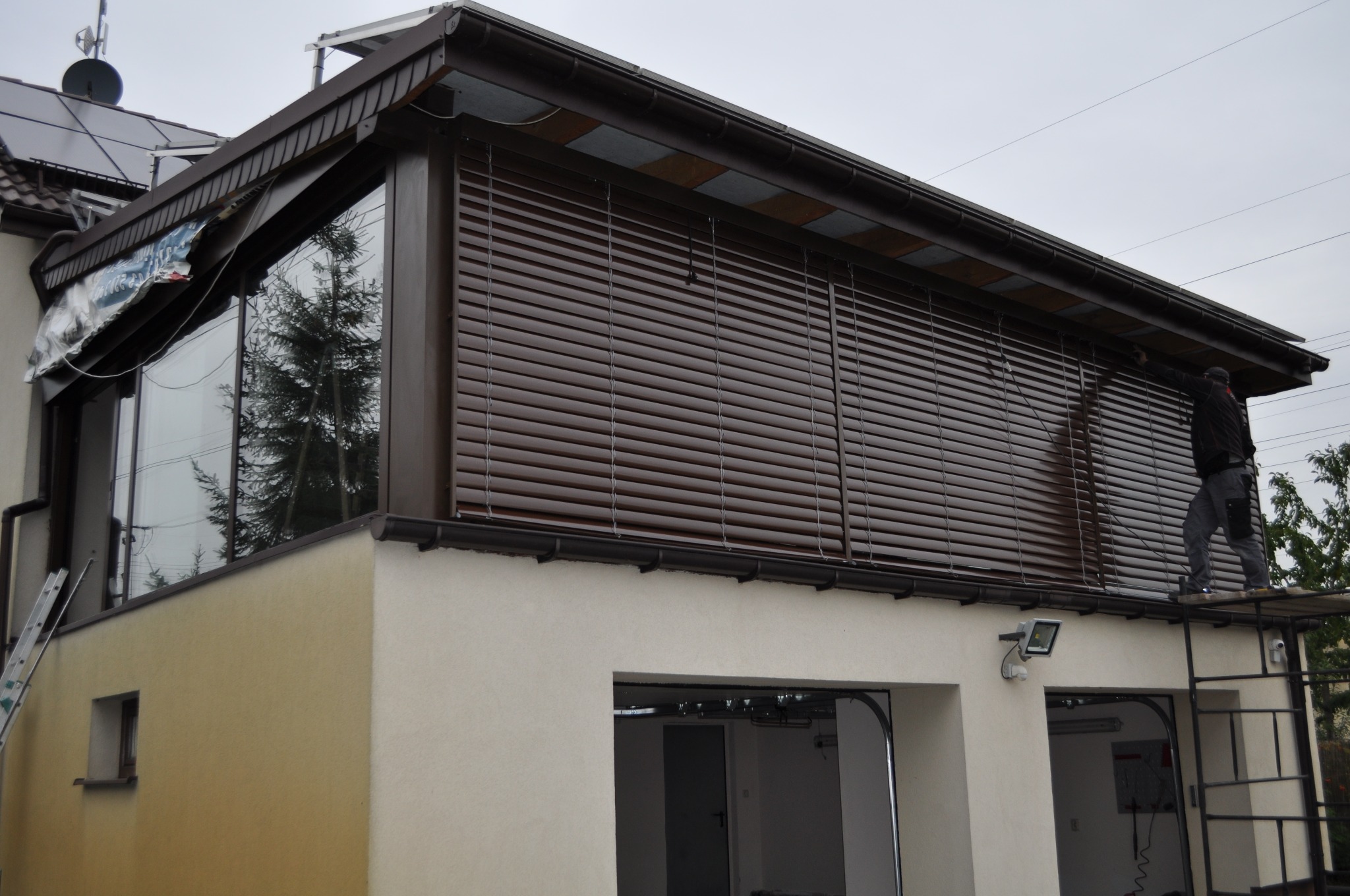 Montaż zewnętrznych, poziomych, brązowych żaluzji na elewacji budynku z widocznymi oknami i garażem, pracownik na rusztowaniu podczas instalacji.