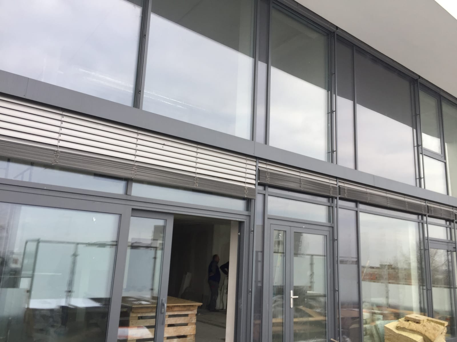 Duże aluminiowe okna fasadowe z żaluzjami zewnętrznymi, widoczne z zewnątrz budynku w trakcie prac wykończeniowych.