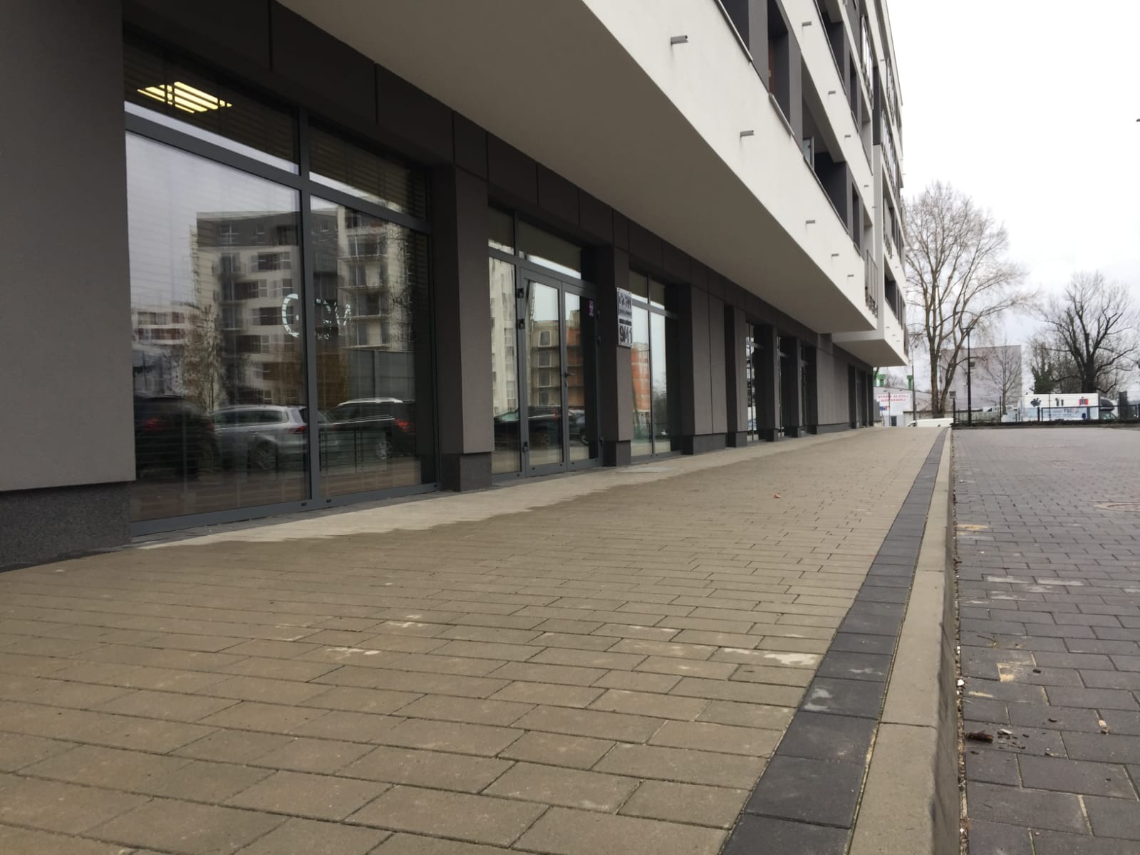 Fasada nowoczesnego budynku biurowego z dużymi, przeszklonymi witrynami i szarymi ramami okiennymi, odbijającymi otoczenie, widok z poziomu chodnika wyłożonego kostką.