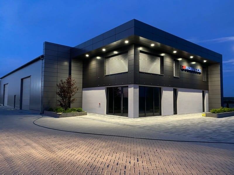 Nowoczesny budynek biurowy z aluminiową elewacją w odcieniach szarości, z oknami z żaluzjami i logo firmy na fasadzie, sfotografowany o zmierzchu.