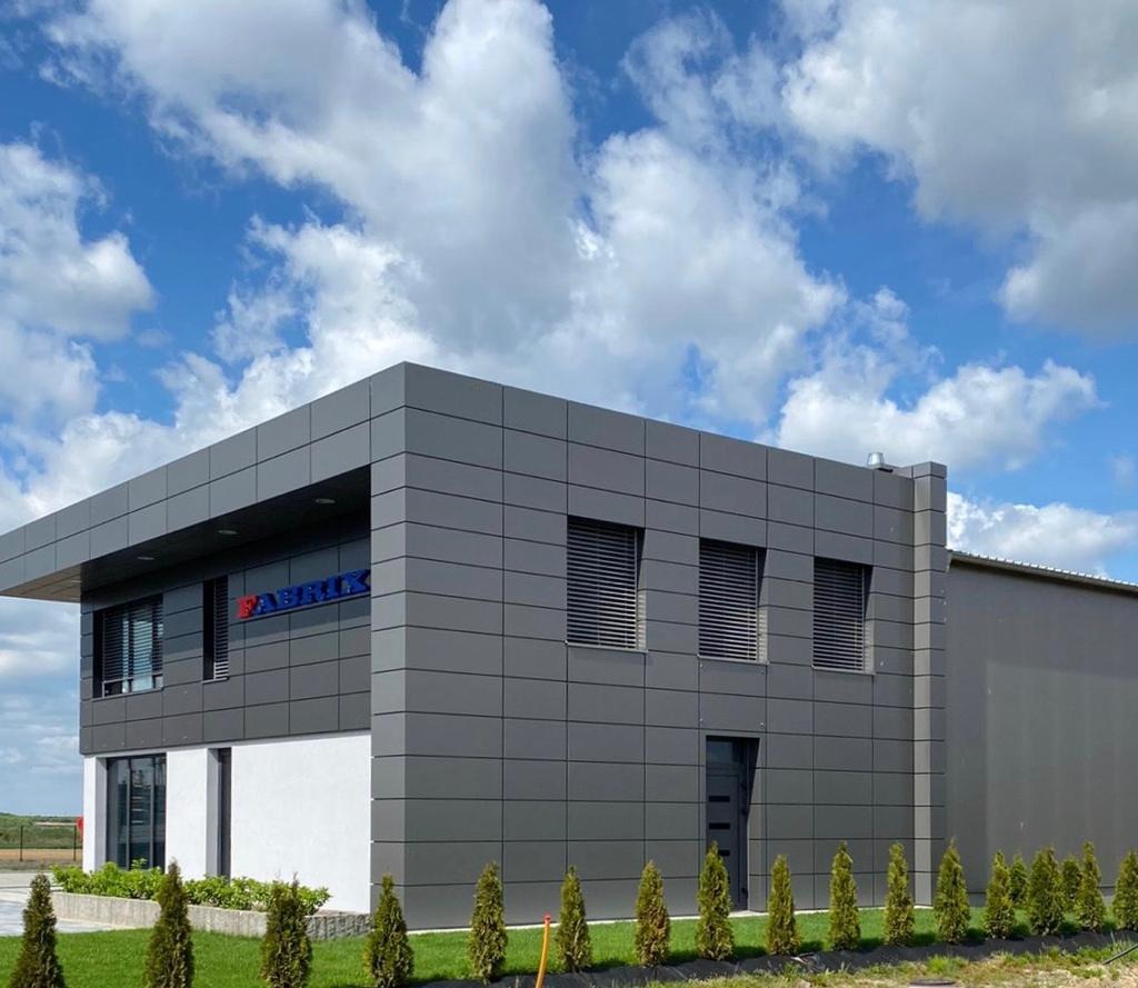 Nowoczesny budynek biurowy z szarą elewacją, oknami z żaluzjami poziomymi i logo firmy 'FABRIX' na fasadzie, otoczony trawnikiem z małymi drzewkami iglastymi, na tle błękitnego nieba z obłokami.