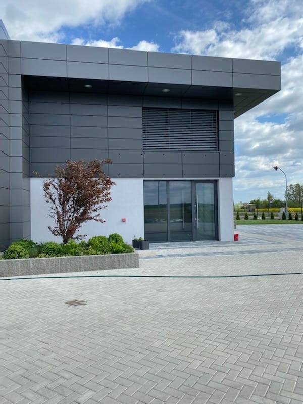 Nowoczesny budynek biurowy z szarą elewacją i aluminiowymi oknami żaluzjowymi, przeszklone wejście, bruk na pierwszym planie, rabata z zielenią i drzewkiem.