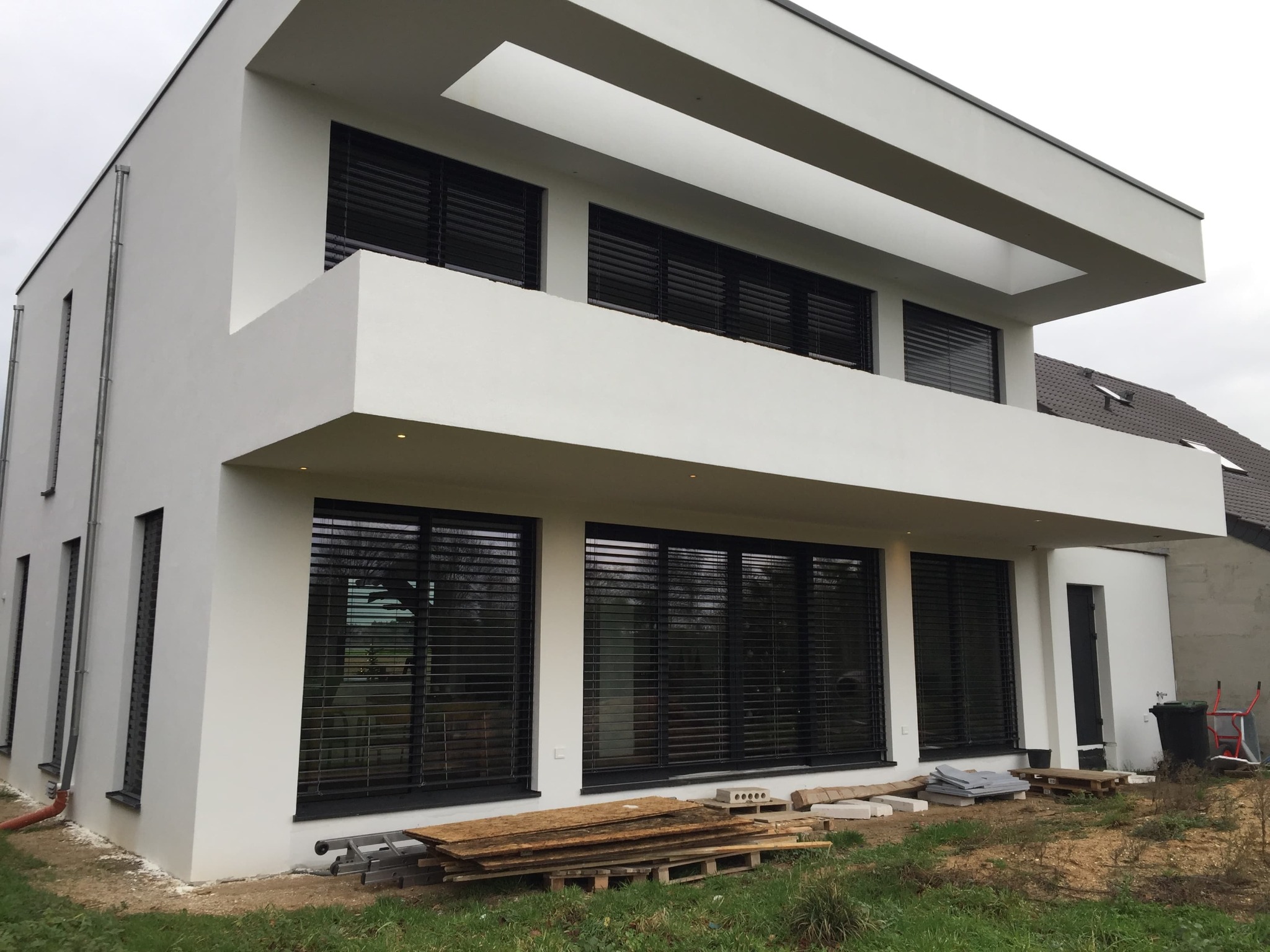 Nowoczesny, dwukondygnacyjny dom z białymi ścianami i dużymi oknami z czarnymi żaluzjami, otoczony trawnikiem i materiałami budowlanymi.