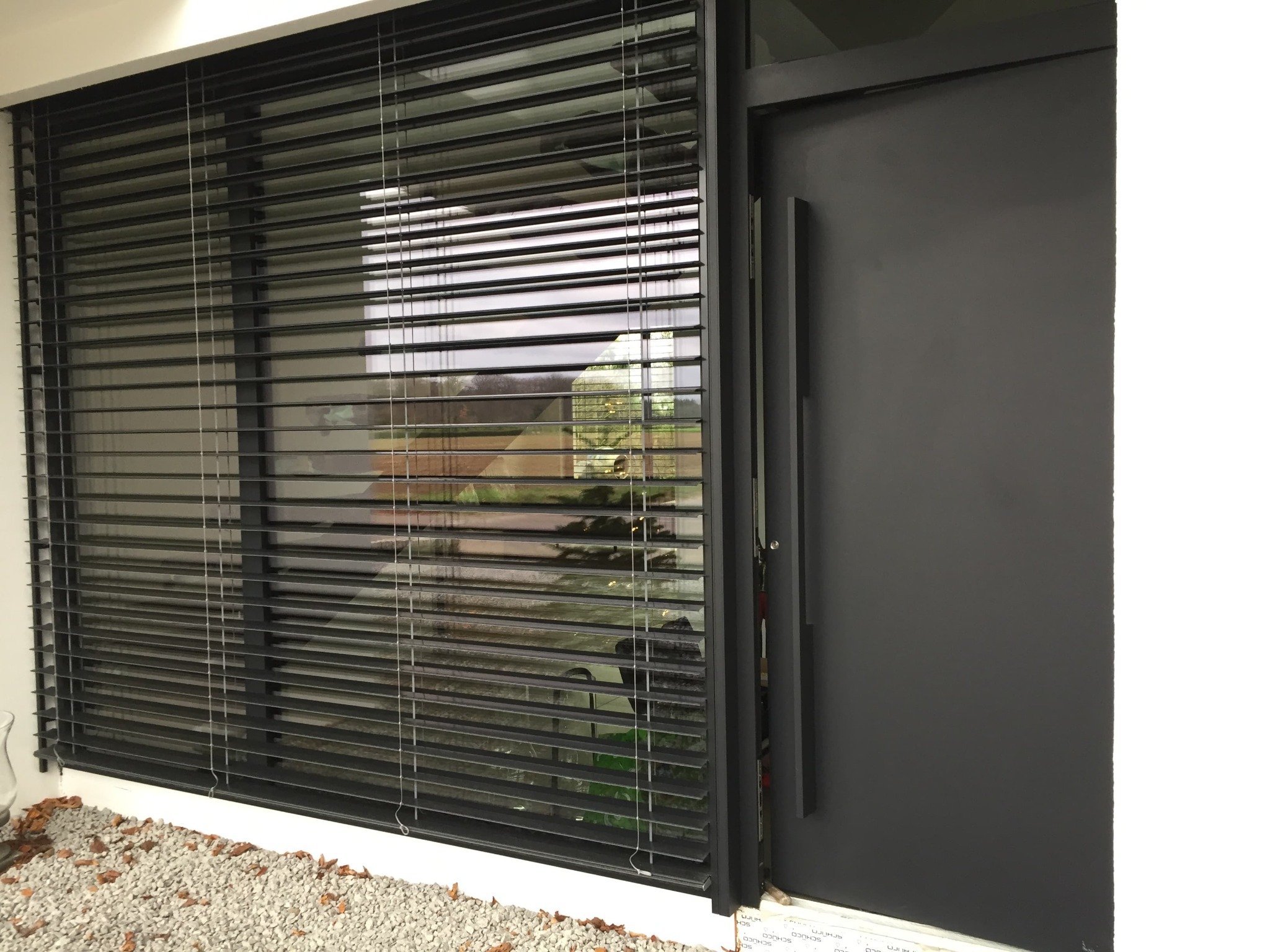 Nowoczesne okno z czarnymi żaluzjami poziomymi, widok z zewnątrz, obok drzwi wejściowe w kolorze antracytowym, jasne otoczenie budynku.