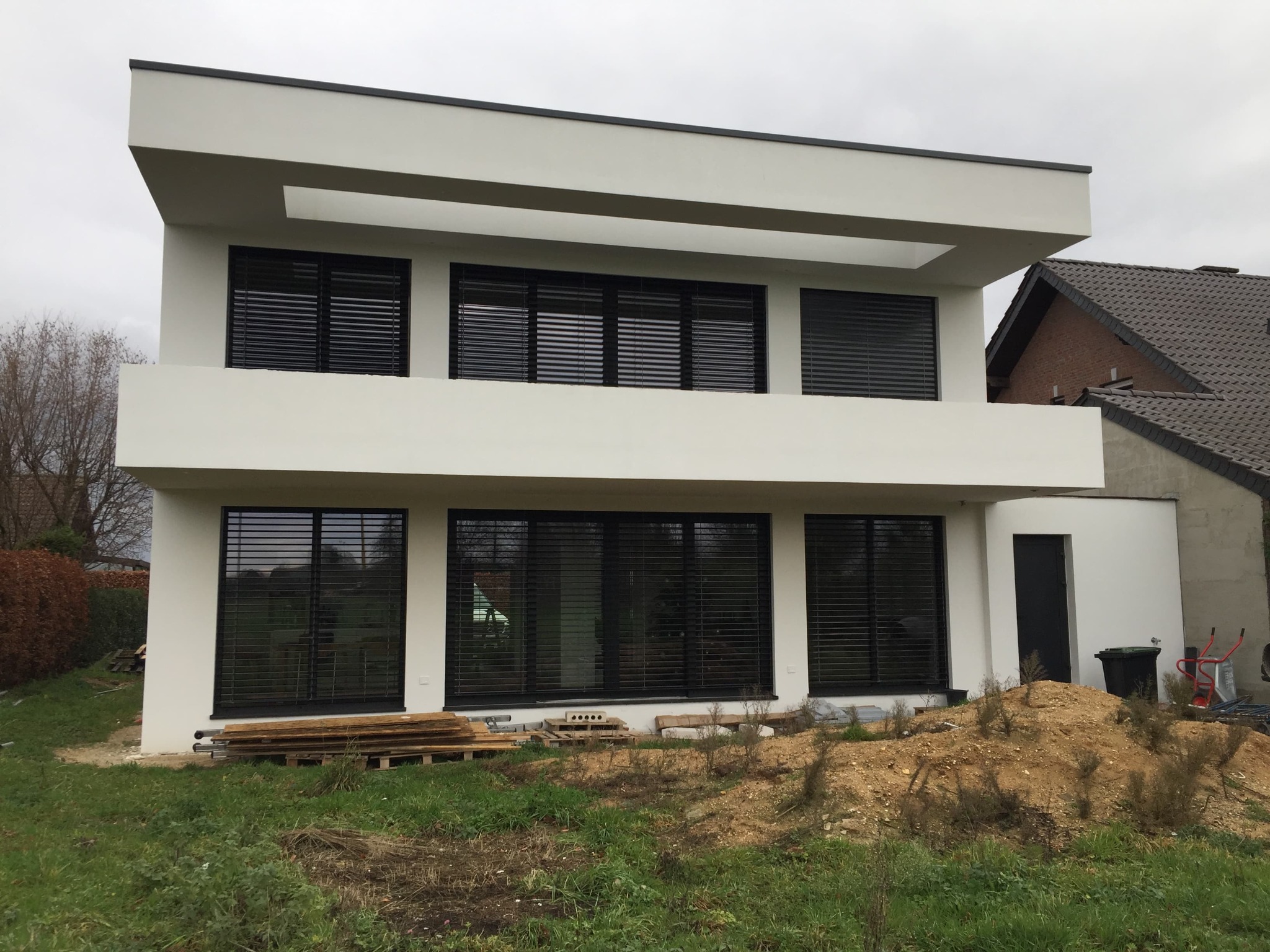 Nowoczesny, dwupiętrowy dom z białymi ścianami i dużymi, czarnymi oknami z żaluzjami, widoczne drewniane deski i narzędzia budowlane na trawniku, sugerujące prace wykończeniowe.