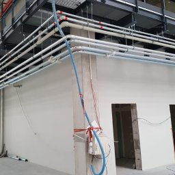 NIKALE Andrzej Maniak - Wnętrze hali przemysłowej w trakcie instalacji rurociągów pod stropem, widoczne liczne rury prowadzone pod sufitem oraz pracownik w oddali.