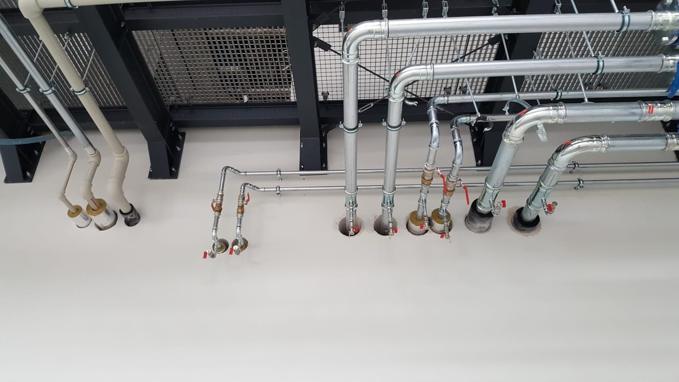 Widok z dołu na instalację rur z zaworami, częściowo chromowanych, przechodzących przez otwory w suficie, z widoczną konstrukcją wsporczą pod stropem.