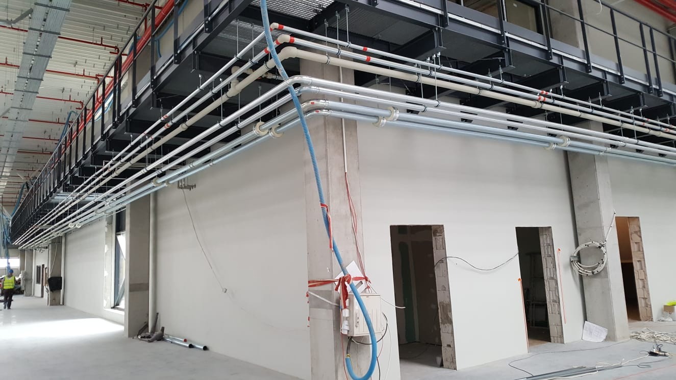 Wnętrze hali przemysłowej w trakcie instalacji rurociągów pod stropem, widoczne liczne rury prowadzone pod sufitem oraz pracownik w oddali.