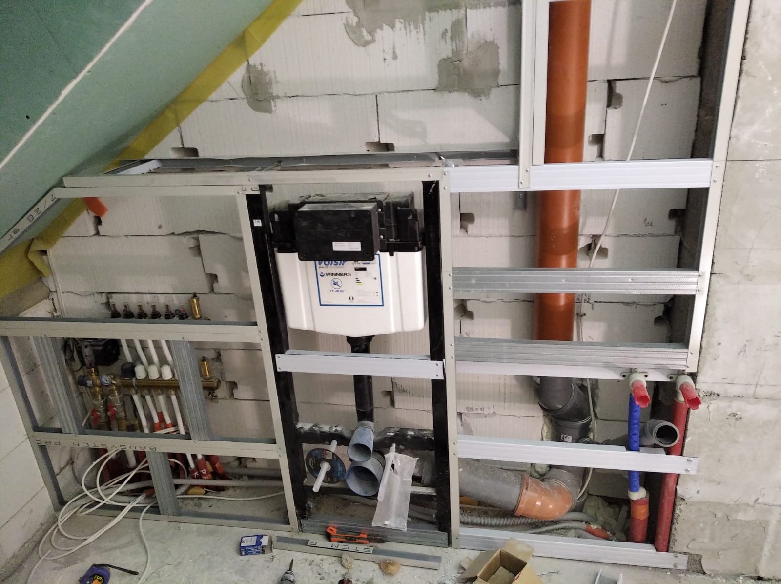 Stelaż podtynkowy WC w trakcie montażu, widoczne rury kanalizacyjne, instalacja wodna z rozdzielaczem, całość w surowym stanie, ściany z betonu komórkowego, poddasze.