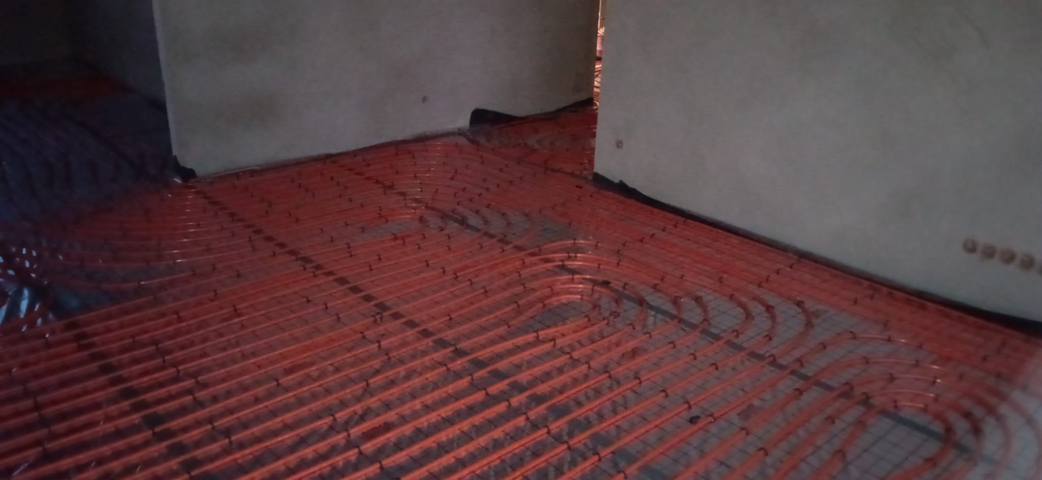 Pomarańczowe rury ogrzewania podłogowego ułożone w pętle na siatce montażowej, widoczne surowe ściany w tle.