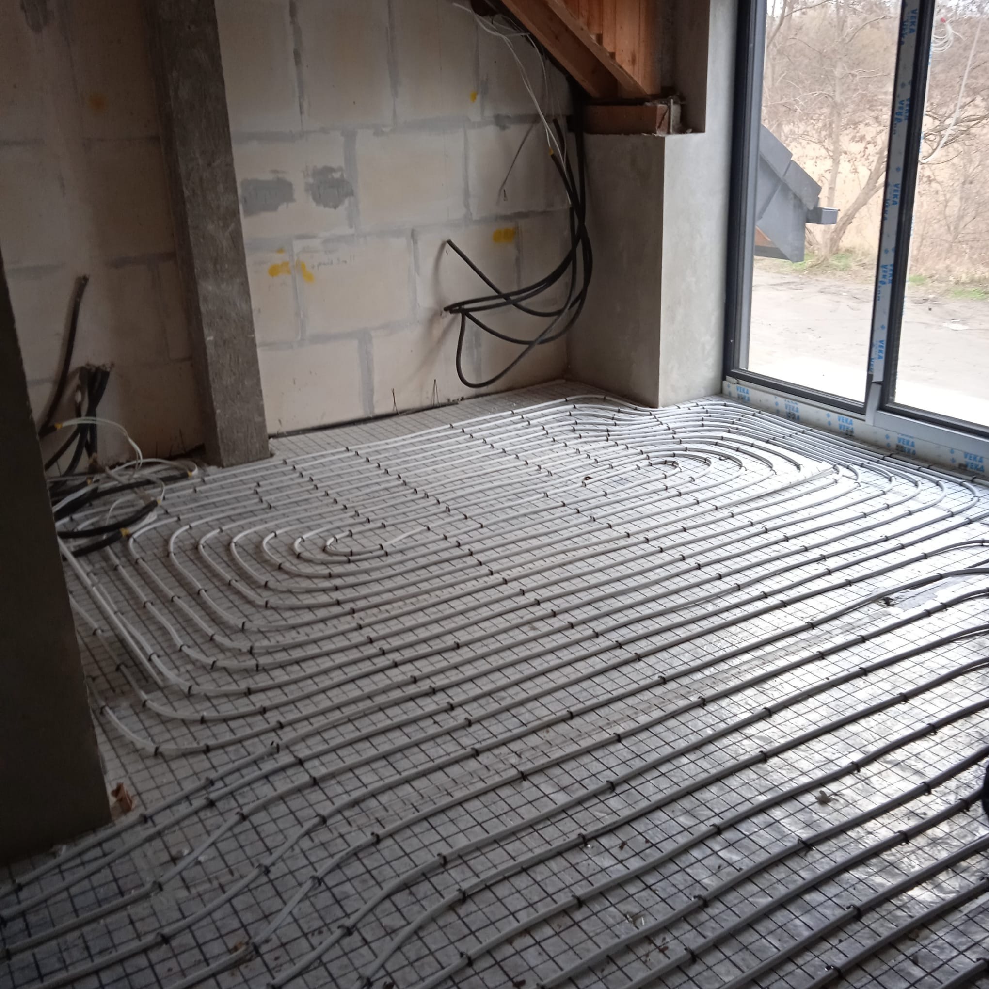 Szare rurki ogrzewania podłogowego ułożone spiralnie na siatce w surowym wnętrzu budynku w trakcie budowy, widoczne kable i okno na zewnątrz.