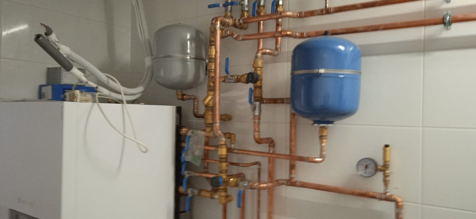 Instalacja grzewcza z kotłem, zbiornikami wyrównawczymi (szarym i niebieskim) oraz miedzianymi rurami i zaworami na tle białych płytek.