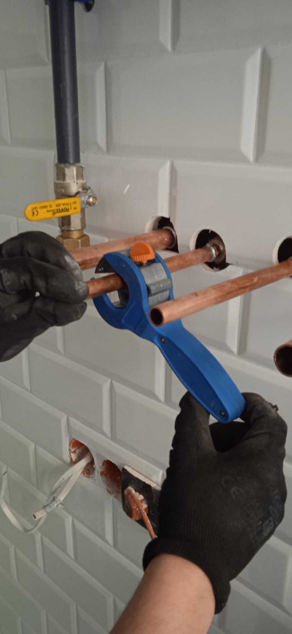 Hydraulik w czarnych rękawicach przycina miedzianą rurę niebieskim obcinakiem na tle białych, kafelkowych ścian z widocznymi przewodami i zaworem.