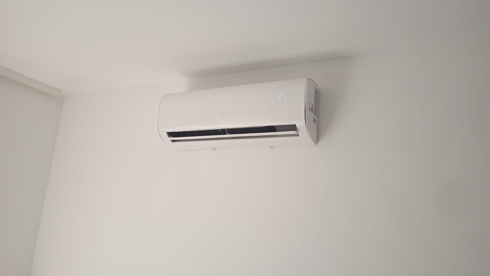 Biały klimatyzator ścienny z wyświetlaczem temperatury 25 stopni, zamontowany na białej ścianie w minimalistycznym wnętrzu.
