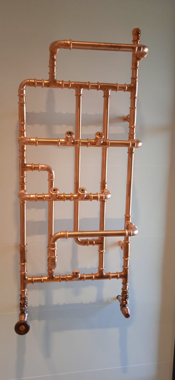 Miedziany grzejnik dekoracyjny w stylu industrialnym zamontowany na ścianie, widoczne liczne złącza i zawory.