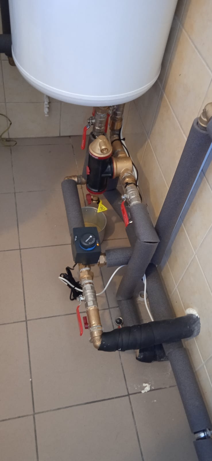 Instalacja grzewcza z kotłem, pompą obiegową i rurami zaizolowanymi szarą pianką, widoczny regulator temperatury i żółty znak ostrzegawczy na wiadrze.