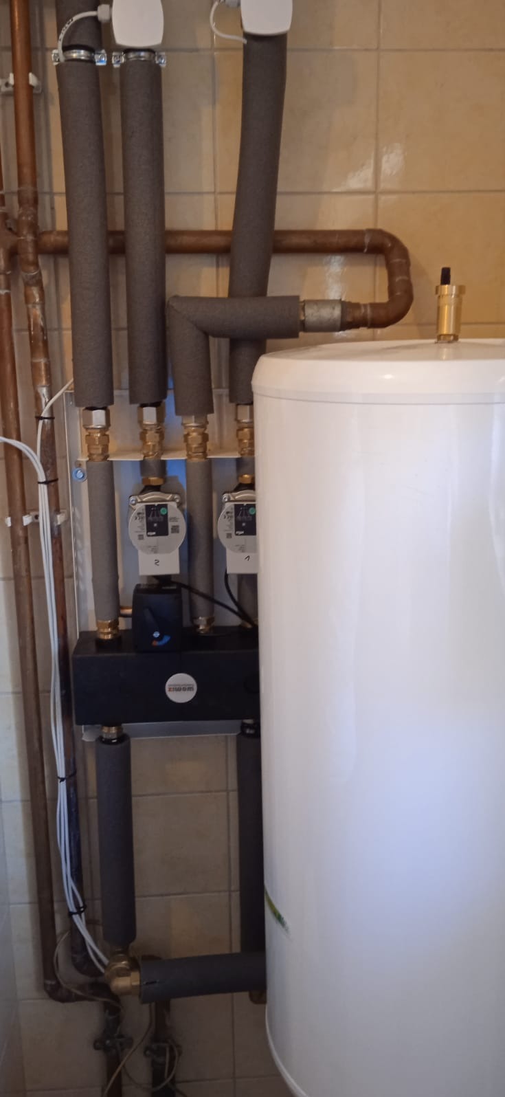 Biały zasobnik ciepłej wody użytkowej z przyłączonymi rurami w izolacji, widoczne pompy obiegowe i armatura, całość na tle wykafelkowanej ściany.