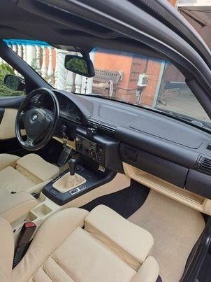 Jasna, skórzana tapicerka wnętrza klasycznego BMW E30 Cabrio, widok z zewnątrz na deskę rozdzielczą, kierownicę i fotele.