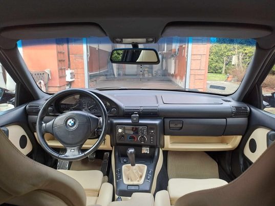 Wnętrze klasycznego BMW E36 z jasną skórzaną tapicerką, widok z tylnej kanapy na deskę rozdzielczą i kierownicę z logo BMW.