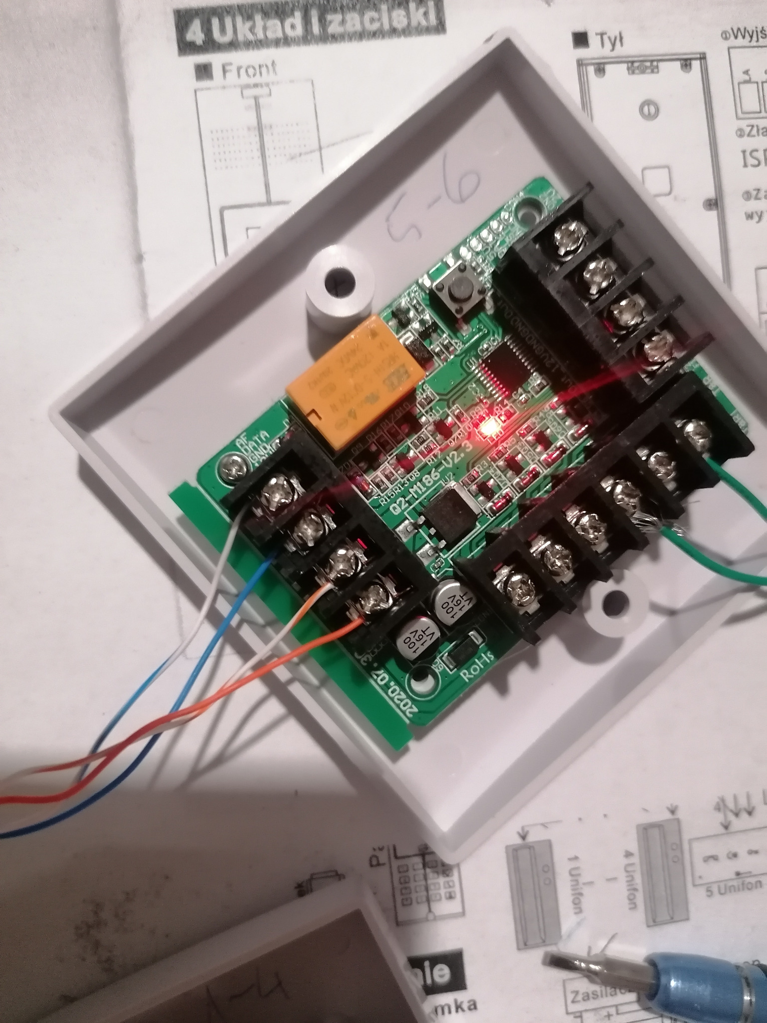 Płyta sterująca z podłączonymi przewodami w białej obudowie, widoczna dioda LED, schemat podłączenia i śrubokręt, w trakcie instalacji.