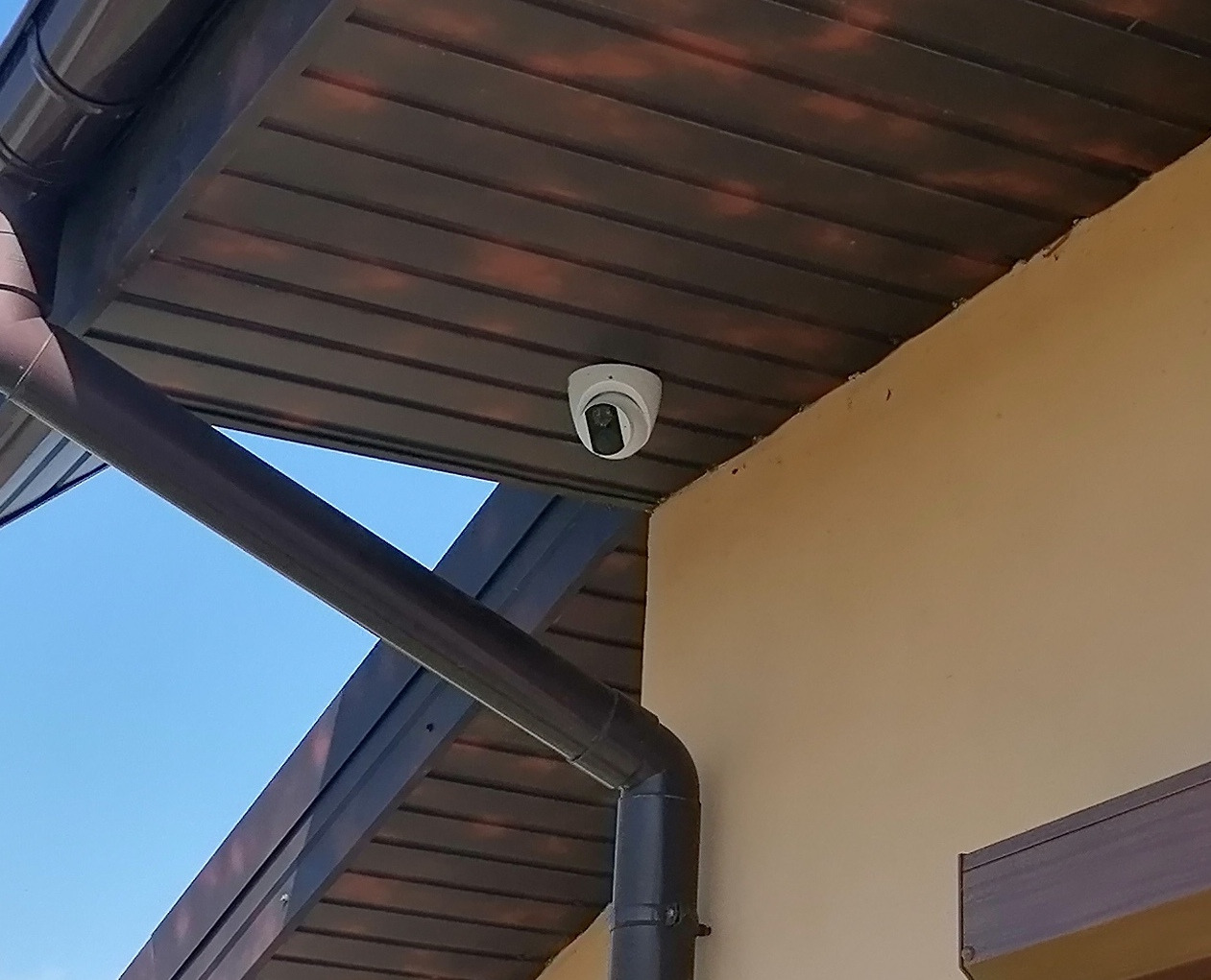 Kamera kopułkowa zamontowana pod okapem budynku z brązową rynną, widok z dołu na tle błękitnego nieba.