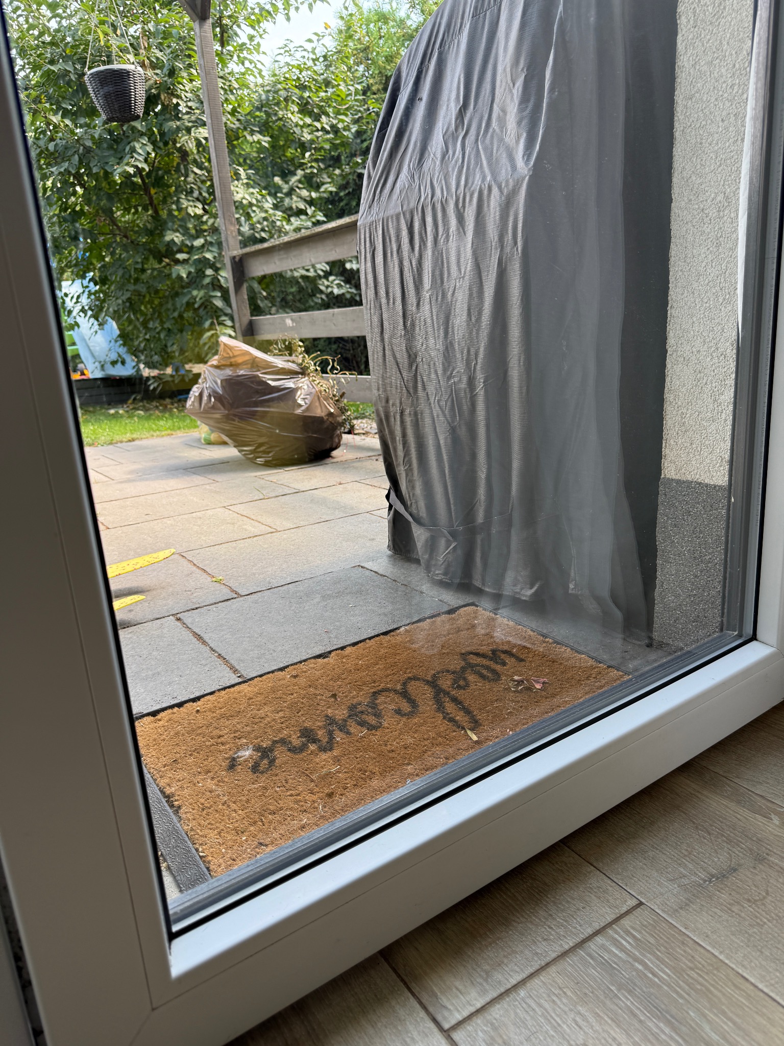 Widok przez okno na taras z wycieraczką 'welcome', workiem śmieci i meblami ogrodowymi pod pokrowcem. Widoczne zabrudzenia na wycieraczce.