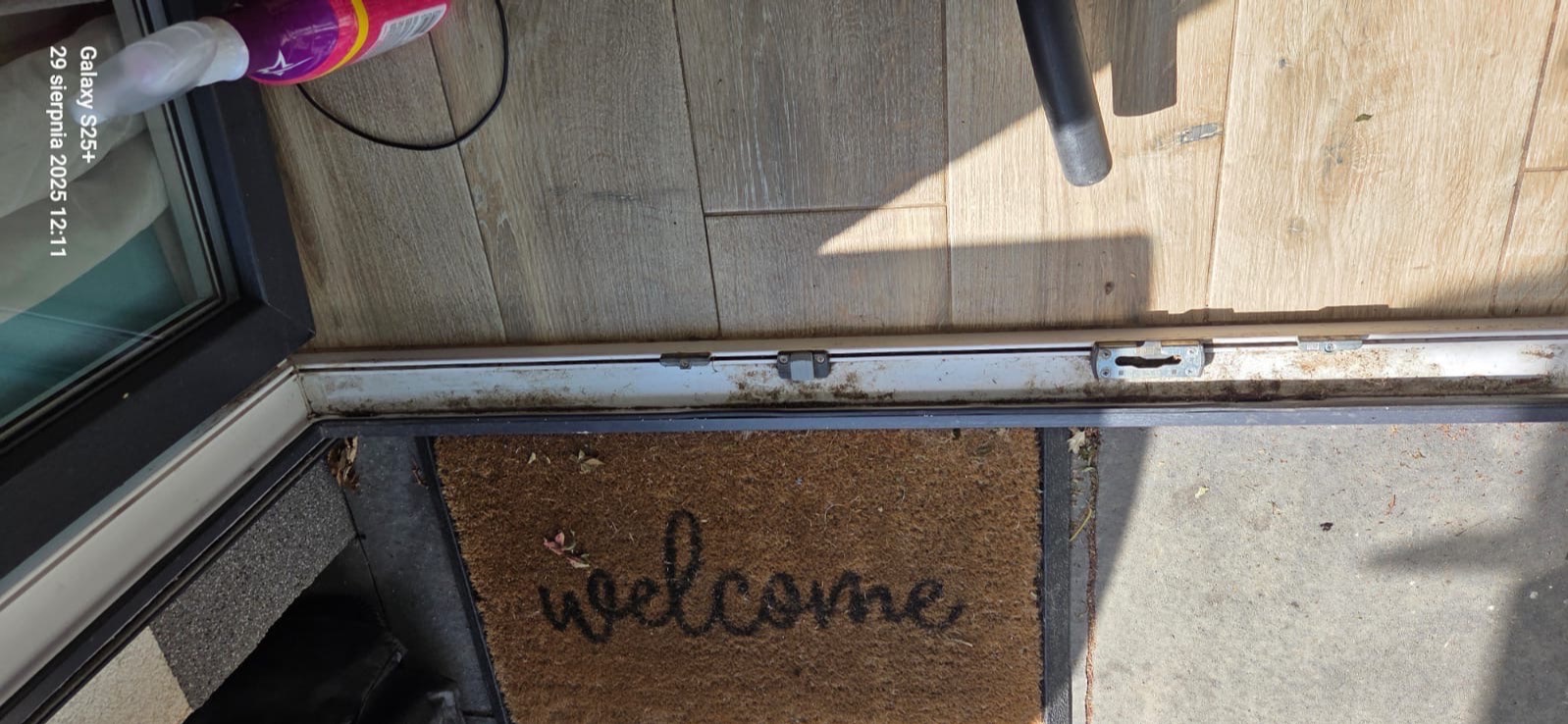 Brudny próg drzwi balkonowych z wycieraczką 'welcome', widoczne ślady kurzu i zabrudzeń na podłodze. Niezadbane detale wymagają dokładnego sprzątania.